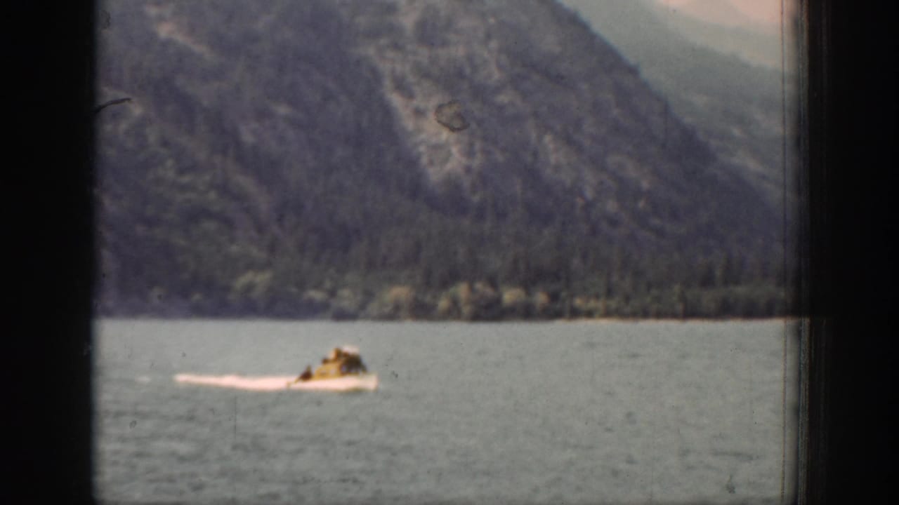 A small boat carrying distant people crosses Lake Chelan, with a white wake and mountain slopes in the background.