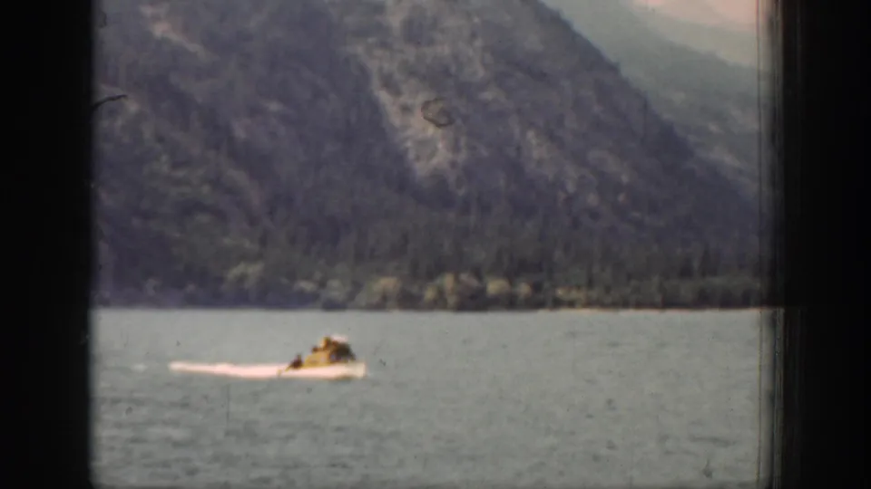 A small boat carrying distant people crosses Lake Chelan, with a white wake and mountain slopes in the background.