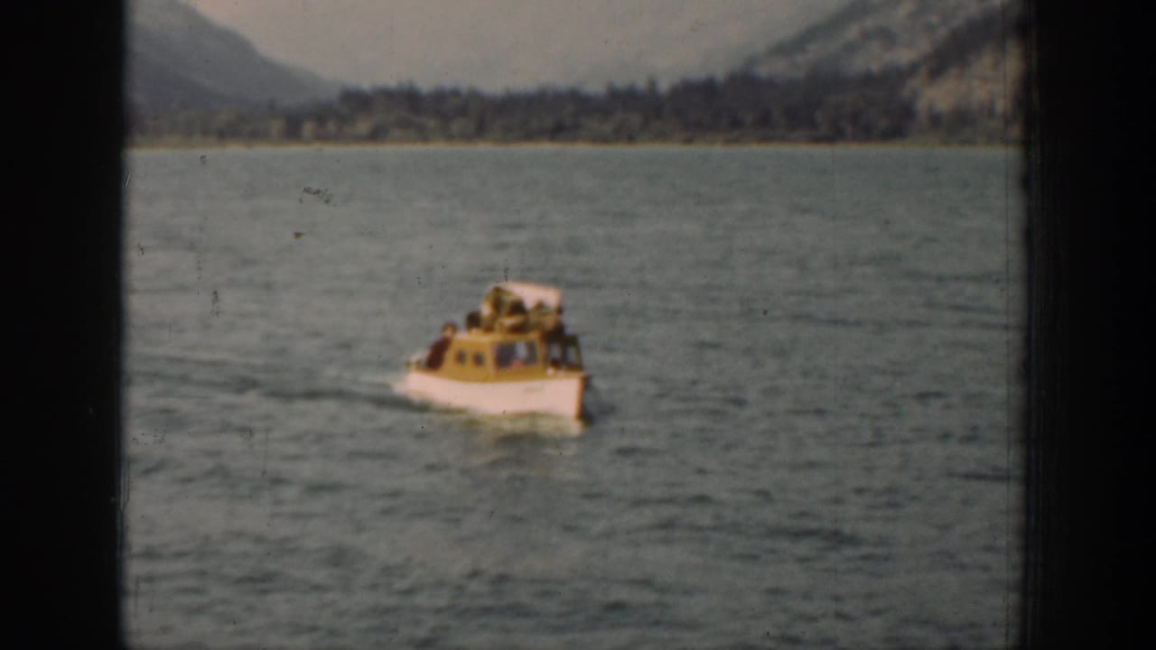 Small cabin motorboat approaches on Lake Chelan, with passengers faintly visible and a mountain shoreline in the distance.