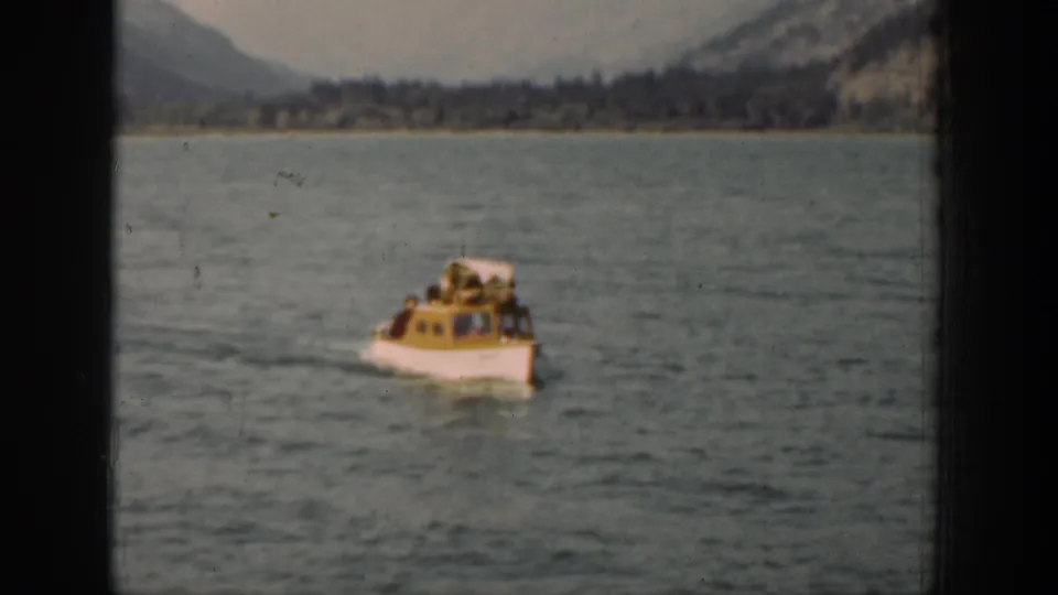 Small cabin motorboat approaches on Lake Chelan, with passengers faintly visible and a mountain shoreline in the distance.