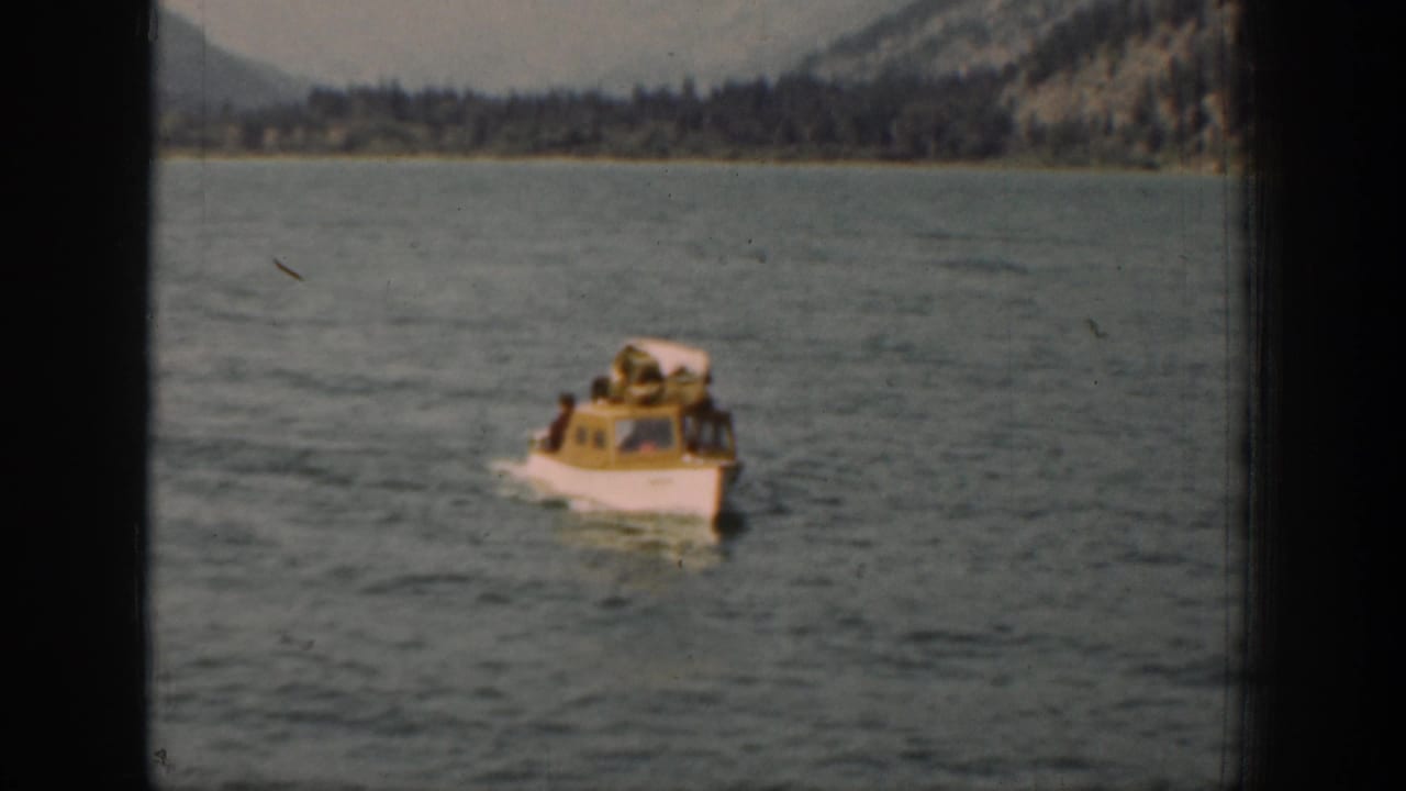 Home-movie view of a small cabin boat on the lake, with passengers visible near the front and wooded hills behind.
