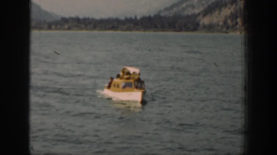 Home-movie view of a small cabin boat on the lake, with passengers visible near the front and wooded hills behind.