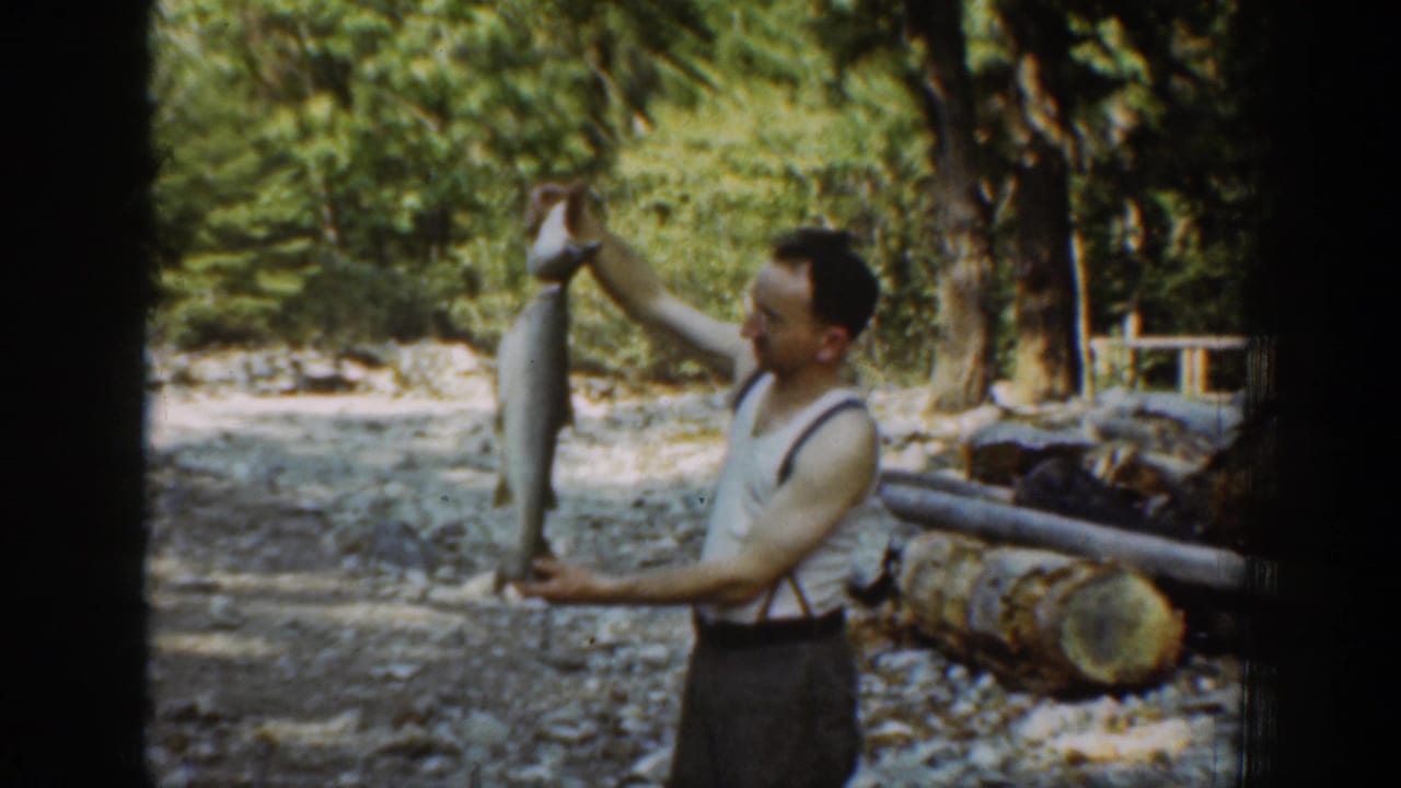 In 1939 Chelan, Washington footage, a man in a sleeveless shirt and suspenders holds up a large fish near logs and trees.