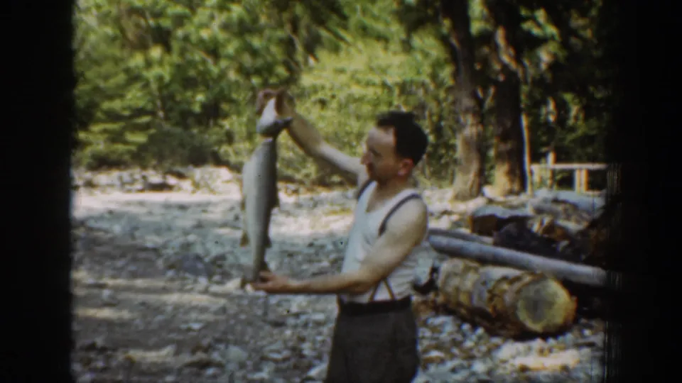 In 1939 Chelan, Washington footage, a man in a sleeveless shirt and suspenders holds up a large fish near logs and trees.