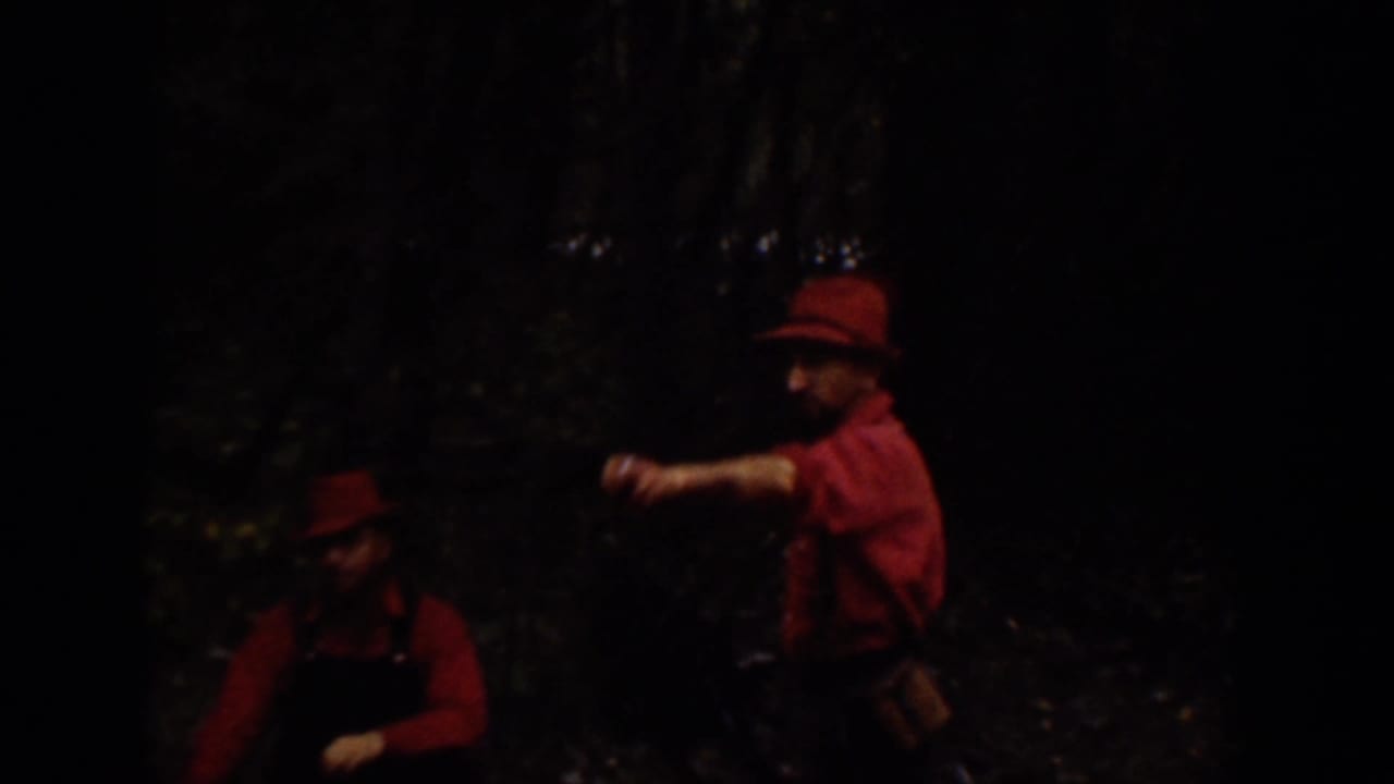 Two men in red shirts and brimmed hats appear in a shaded wooded area, one gesturing while the other stands nearby.