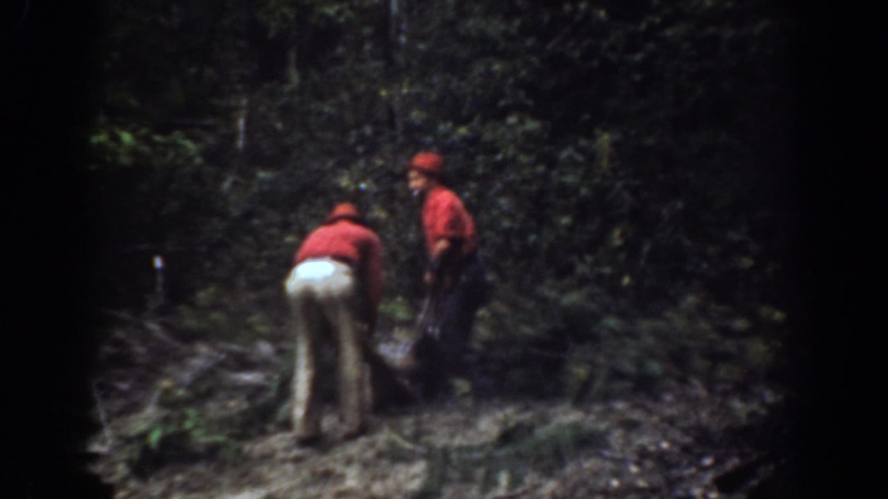 Two men stand and bend near a dark sled or load in a wooded area.