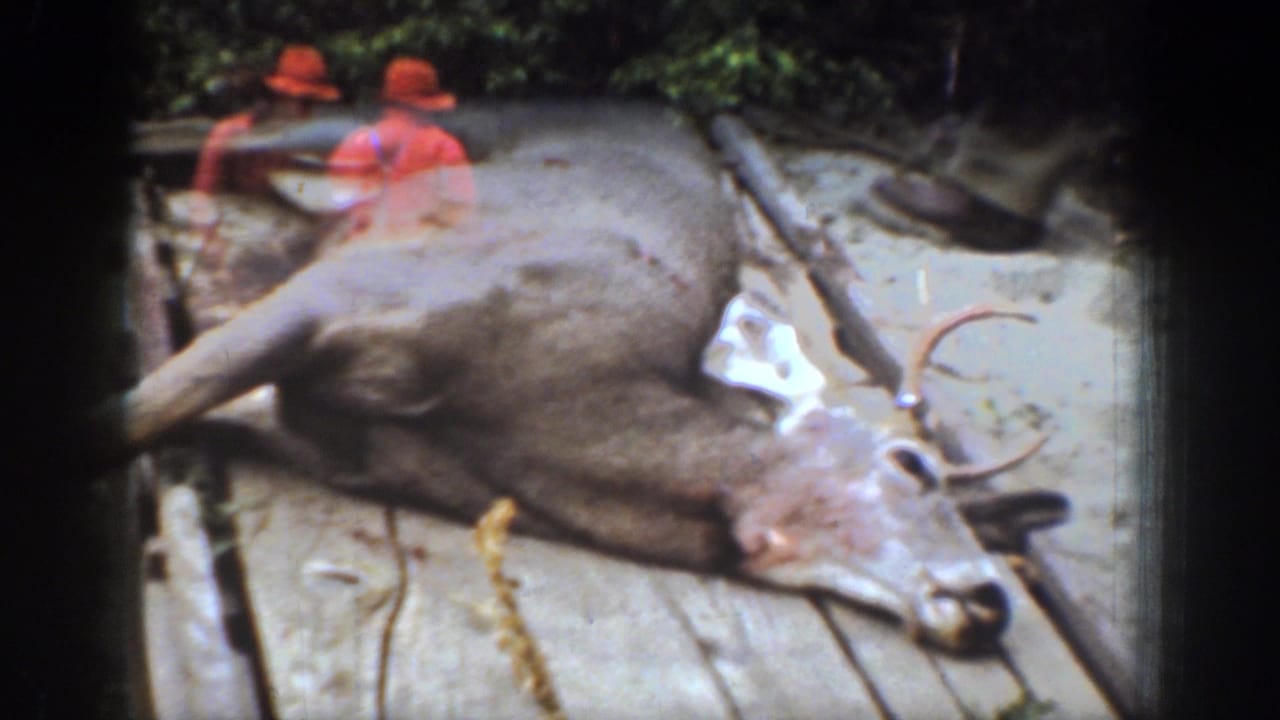 1939 Chelan, Washington home-movie frame showing a harvested antlered deer on a wooden sled with two hunters in red hats nearby.