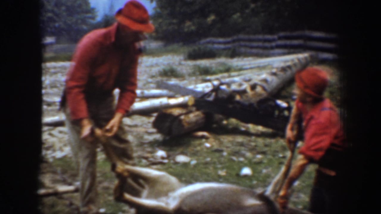 Two men in red hunting clothes stand beside a harvested deer near logs and a sled in the 1939 Chelan, Washington home-movie scene.