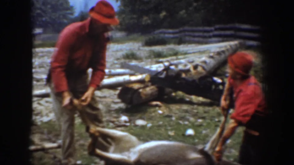 Two men in red hunting clothes stand beside a harvested deer near logs and a sled in the 1939 Chelan, Washington home-movie scene.