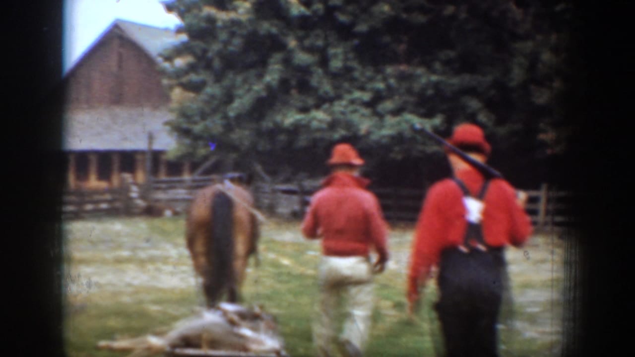 In Chelan, Washington in 1939, men in red hunting clothes walk behind a horse and sled near a wooden barn and fence.