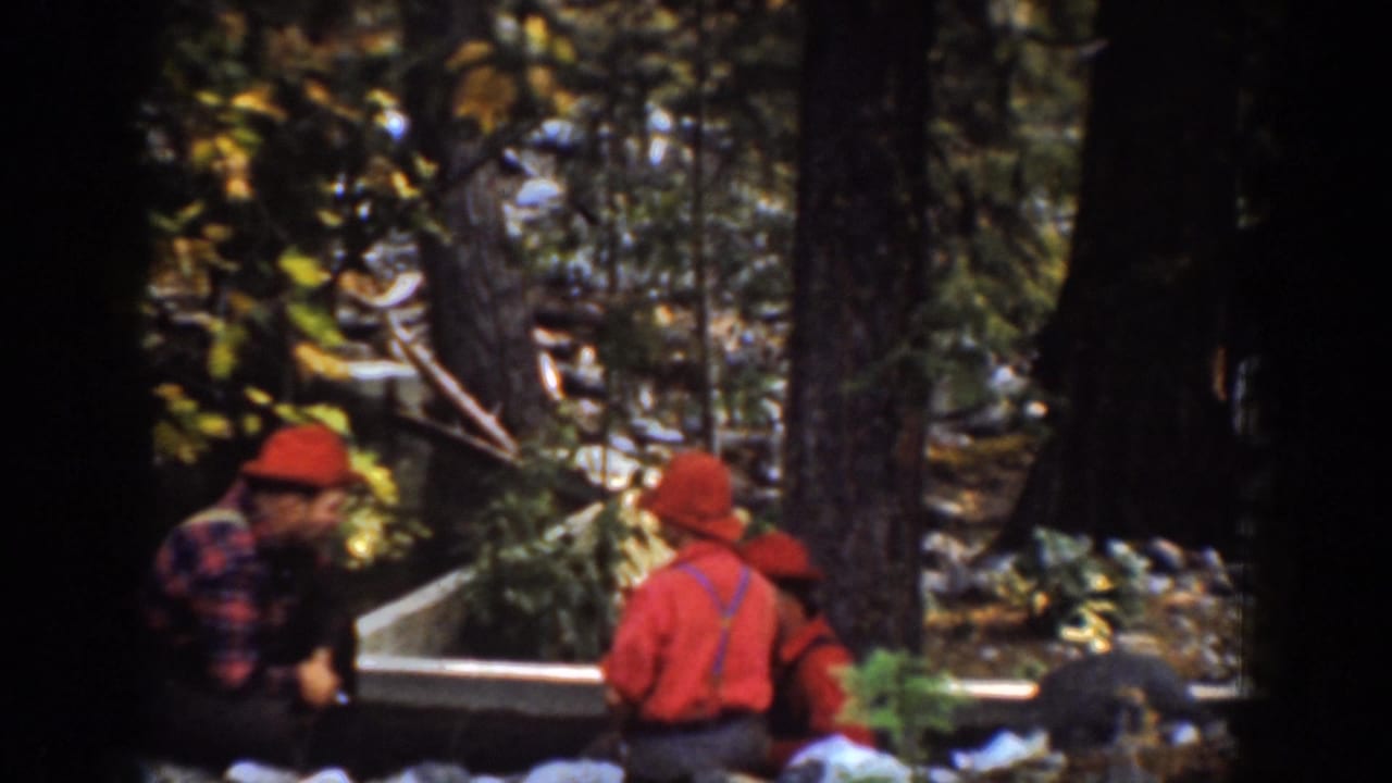 In the Chelan, Washington reel, men in red hats and work clothes gather among trees near a wooden edge or log structure.