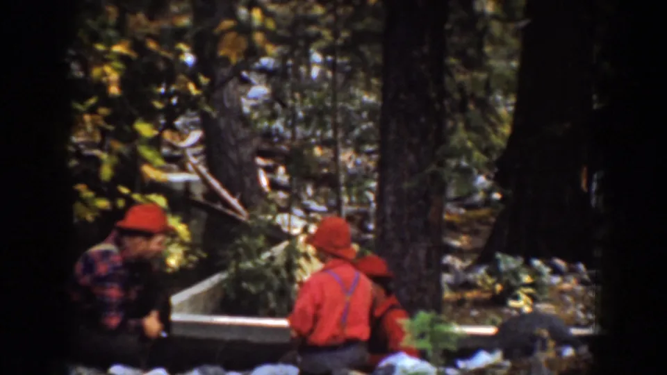 In the Chelan, Washington reel, men in red hats and work clothes gather among trees near a wooden edge or log structure.