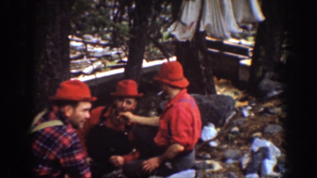 Chelan-area home-movie frame showing men in red hats and work clothes conversing in a forested logging setting.