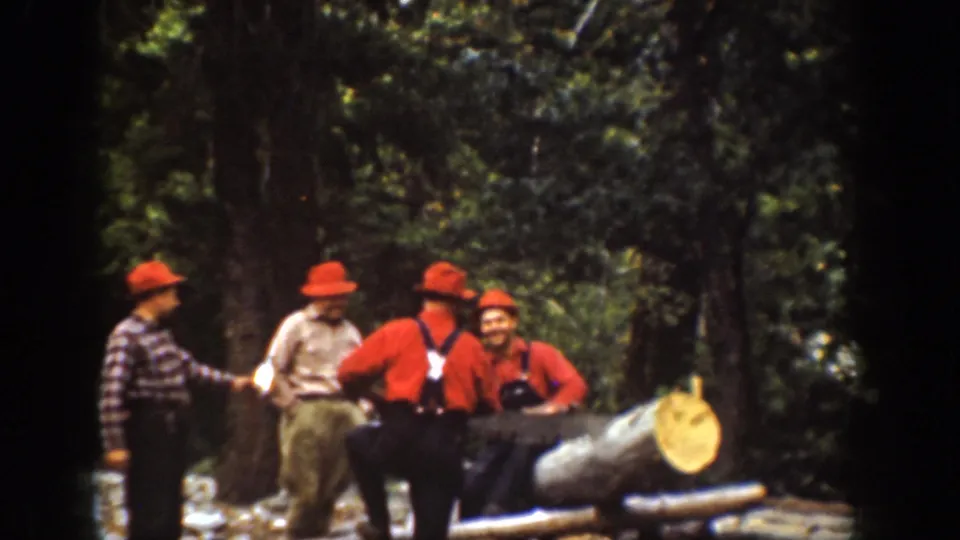 1939 home-movie frame from Chelan showing four men in logging clothes gathered near a large felled tree in the woods.