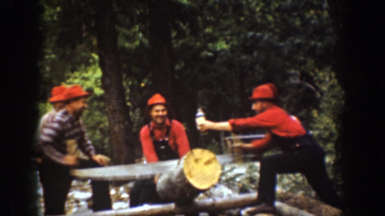 Three men in red hats stand by a large saw and log in a wooded clearing.