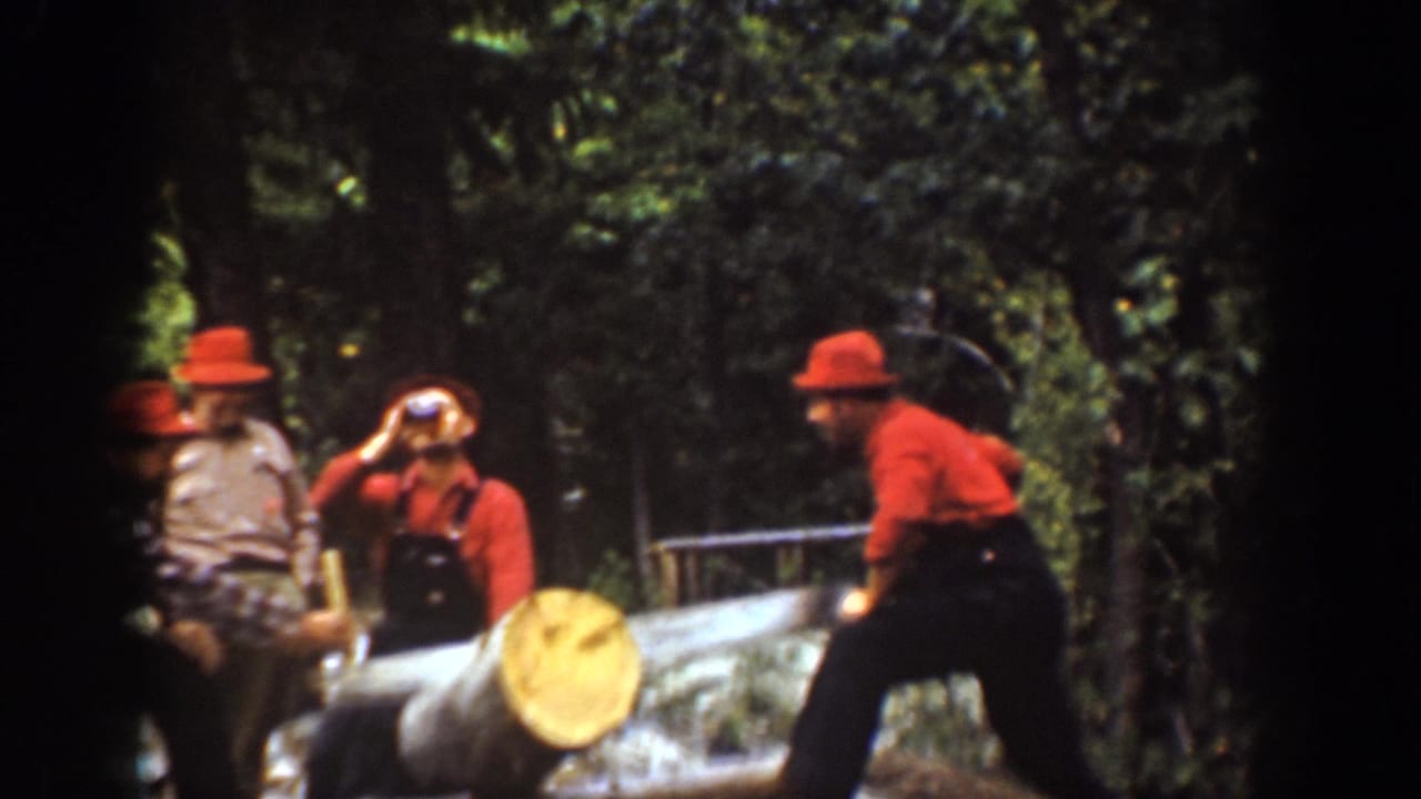 Men stand and sit near a large log while one man raises a bottle and another works beside the log.