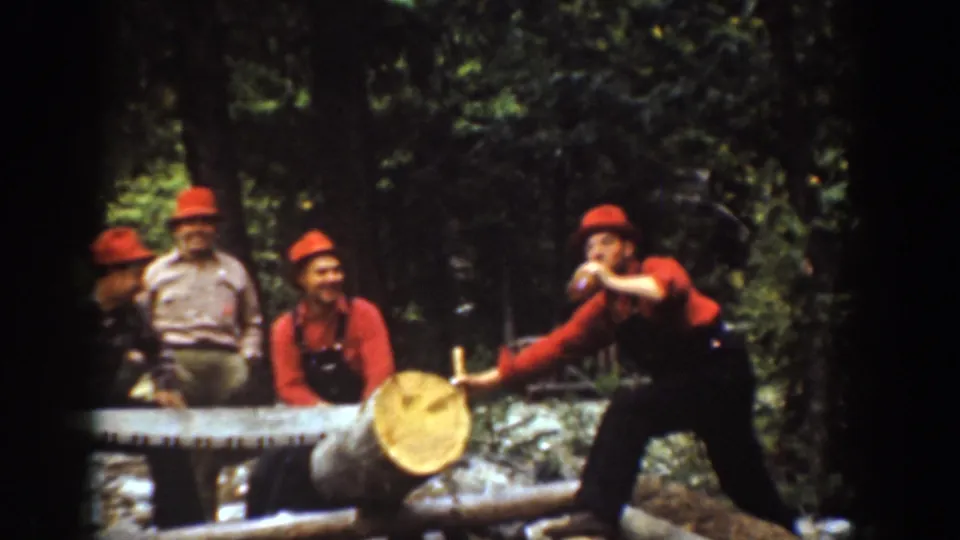 1939 Chelan, Washington home-movie frame showing men around a forest log, with one man extending a bottle near the work area.