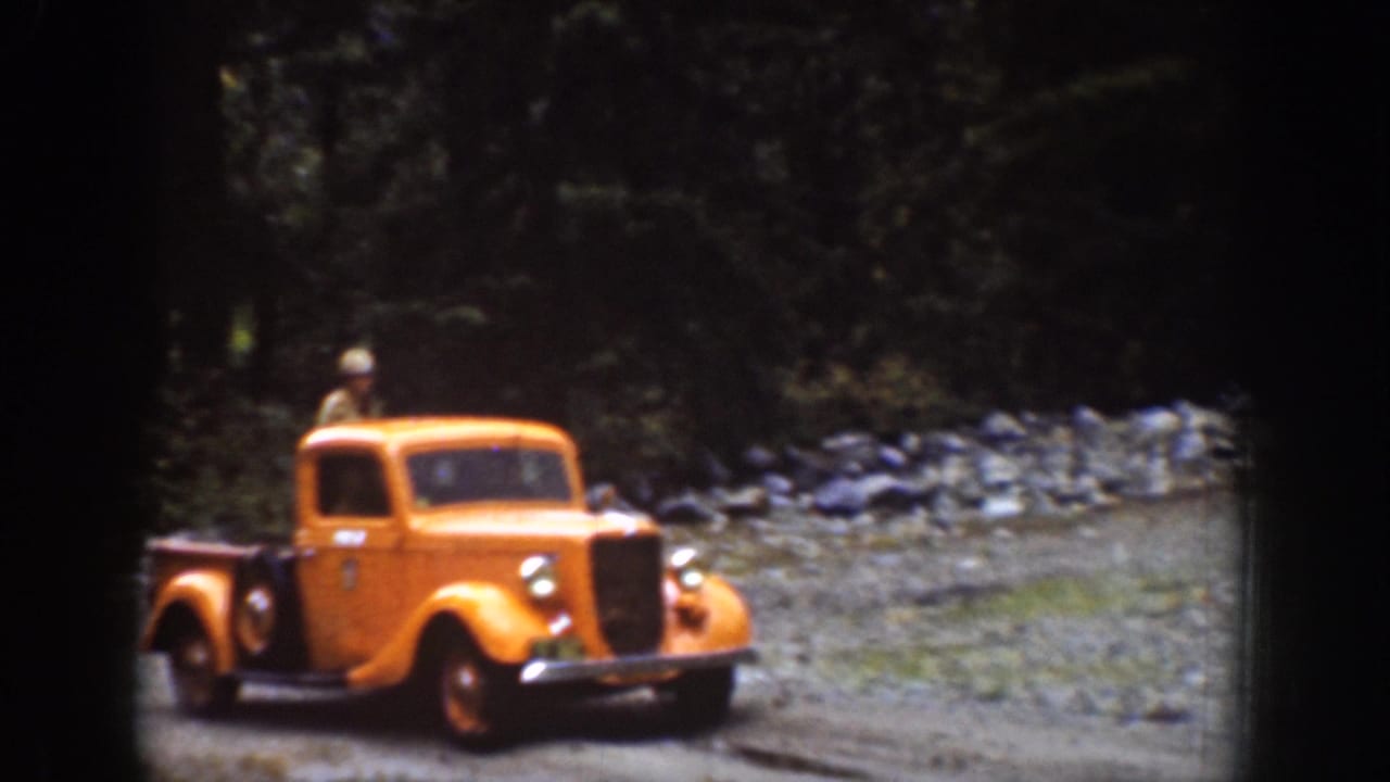 Orange vintage pickup truck driving on a rocky dirt road in a wooded mountain setting.