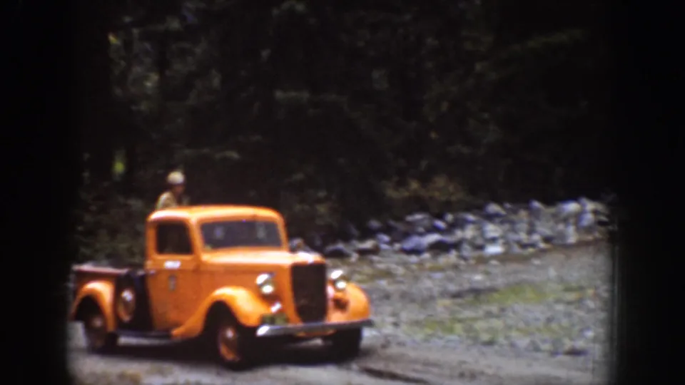 1939 Chelan, Washington home-movie frame showing an orange pickup moving along a rugged dirt road near forest and rock.