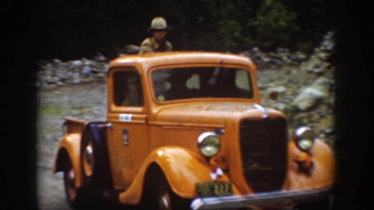 Closer view of orange pickup with a man standing in the truck bed near a rocky creek bed.