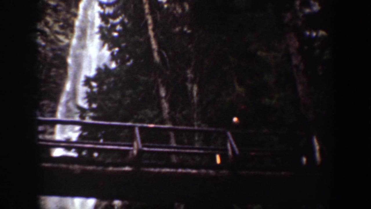 1939 Chelan, Washington 8mm frame showing a bright waterfall behind a wooden observation bridge and dense trees.
