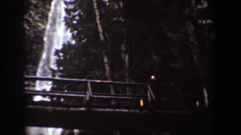 1939 Chelan, Washington 8mm frame showing a bright waterfall behind a wooden observation bridge and dense trees.