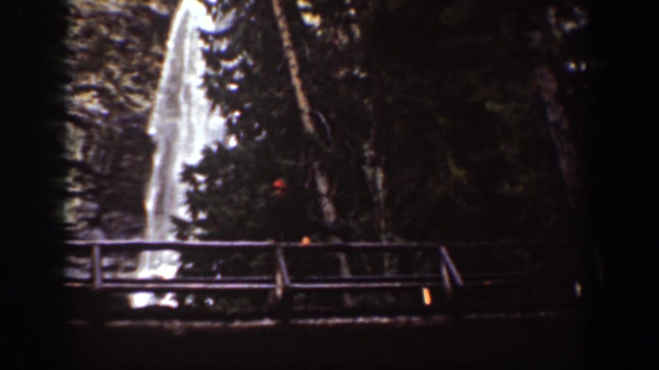 1939 8mm view of a bright cascading waterfall partly obscured by trees, with a bridge railing below.