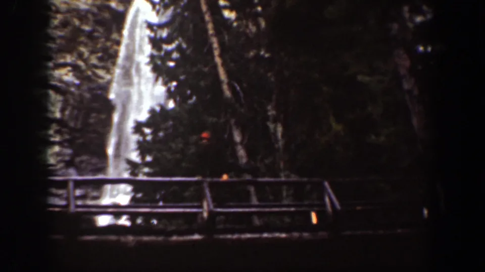 1939 8mm view of a bright cascading waterfall partly obscured by trees, with a bridge railing below.
