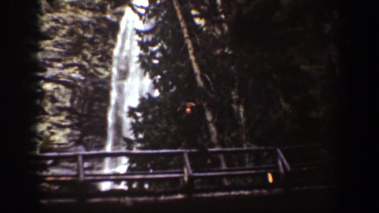 Chelan, Washington 1939 frame showing a tall waterfall, shaded evergreens, rock face, and wooden observation bridge.