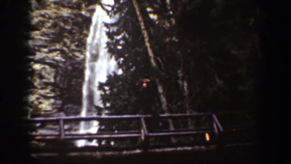 Chelan, Washington 1939 frame showing a tall waterfall, shaded evergreens, rock face, and wooden observation bridge.