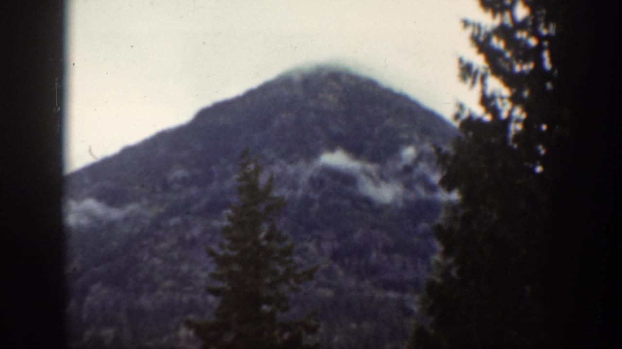 Vintage 8mm frame of a rounded mountain peak near Chelan, with dark conifers and soft low cloud across the slope.