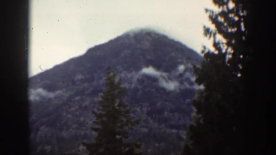 Vintage 8mm frame of a rounded mountain peak near Chelan, with dark conifers and soft low cloud across the slope.