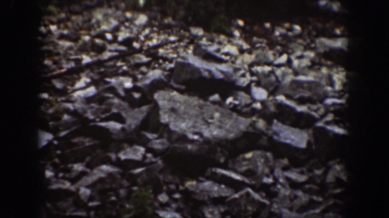 1939 Chelan home-movie frame showing loose dark granite rocks and fractured quarry stone in uneven light.