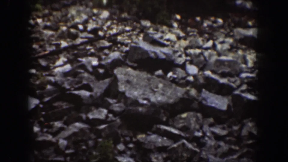 1939 Chelan home-movie frame showing loose dark granite rocks and fractured quarry stone in uneven light.