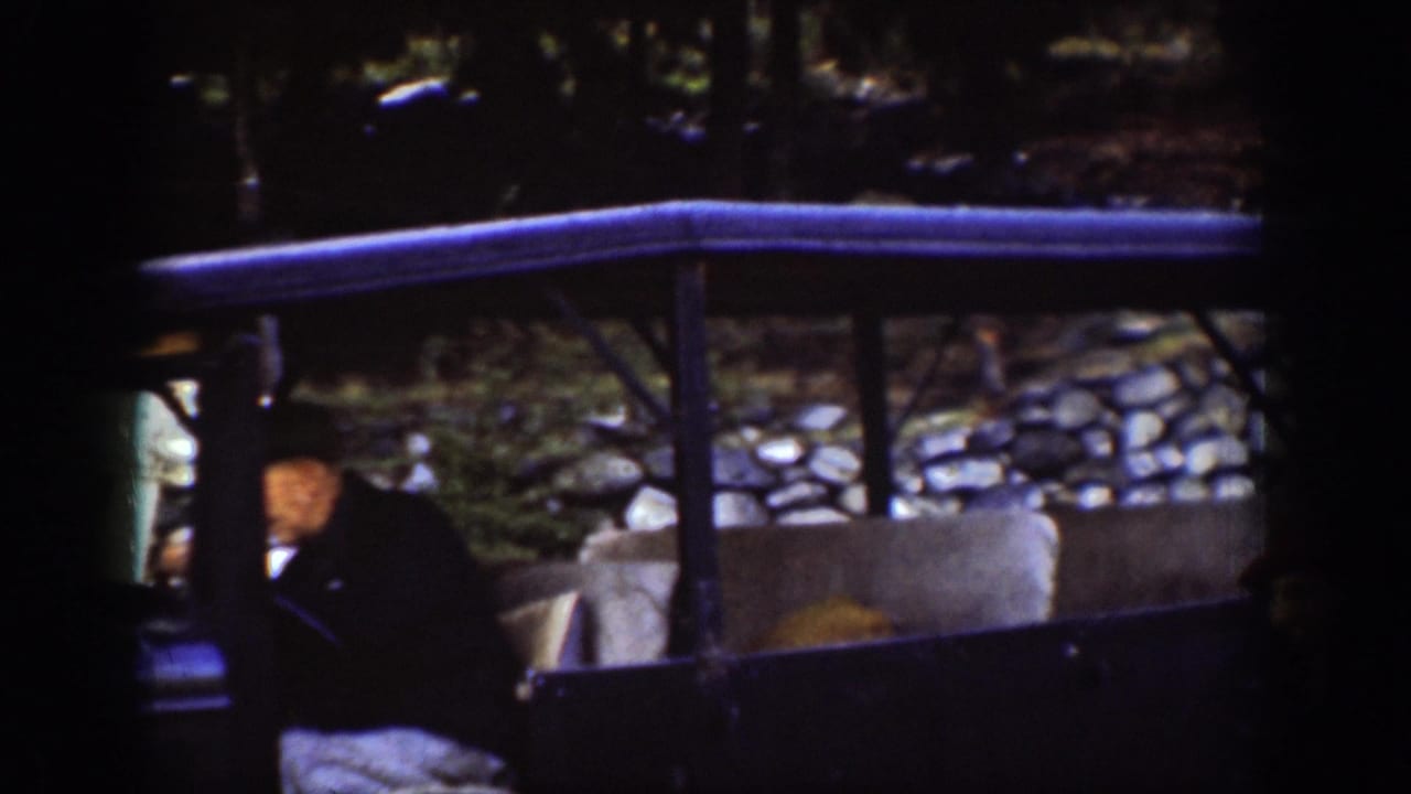 Side view of a vintage vehicle with a wood-framed canopy alongside a stone retaining wall; figures in period coats visible, consistent with a 1939 ferry embarkation point.