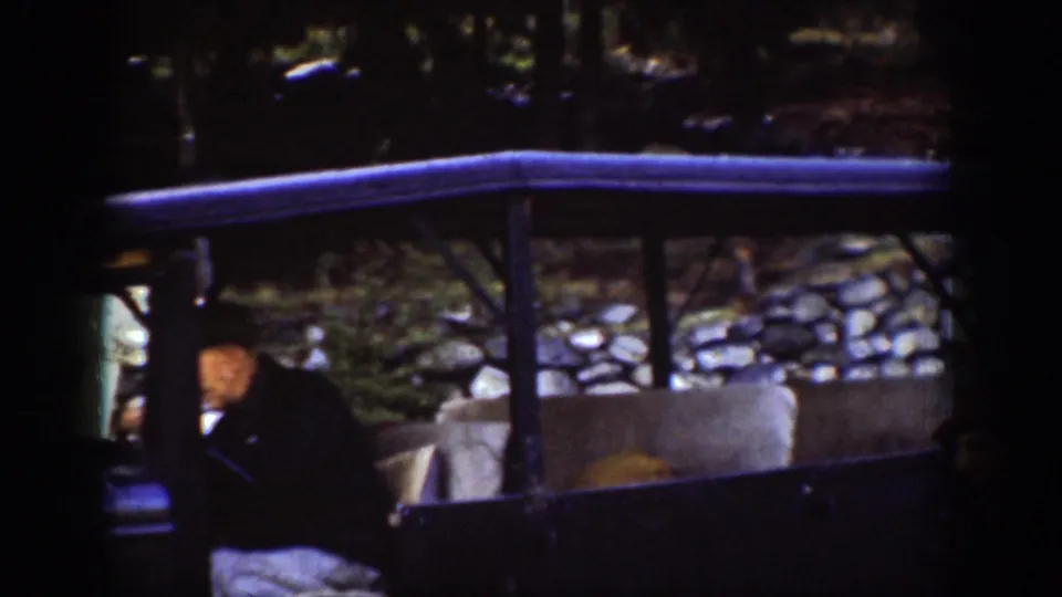 Side view of a vintage vehicle with a wood-framed canopy alongside a stone retaining wall; figures in period coats visible, consistent with a 1939 ferry embarkation point.