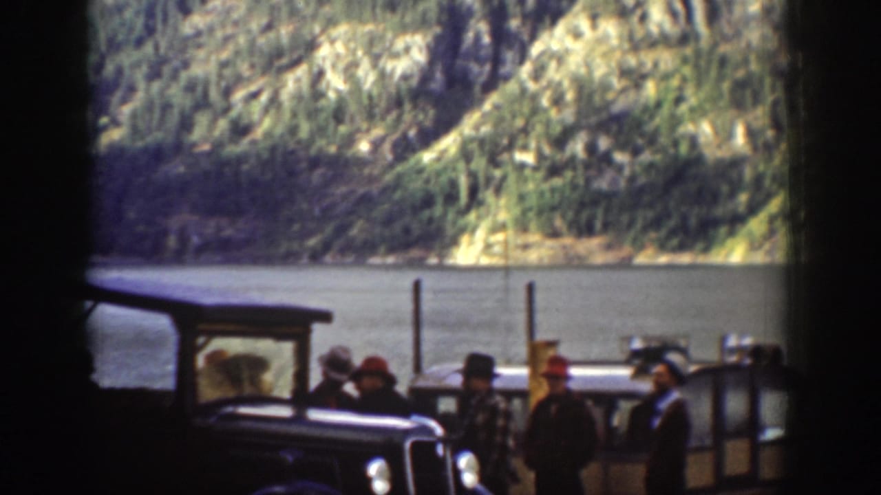 Group in 1930s-era hats and coats gathered near a vintage automobile at an apparent ferry landing; rocky forested hillside and calm lake surface visible, c. 1939 Chelan, Washington.