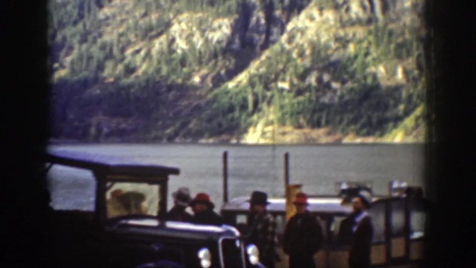 Group in 1930s-era hats and coats gathered near a vintage automobile at an apparent ferry landing; rocky forested hillside and calm lake surface visible, c. 1939 Chelan, Washington.