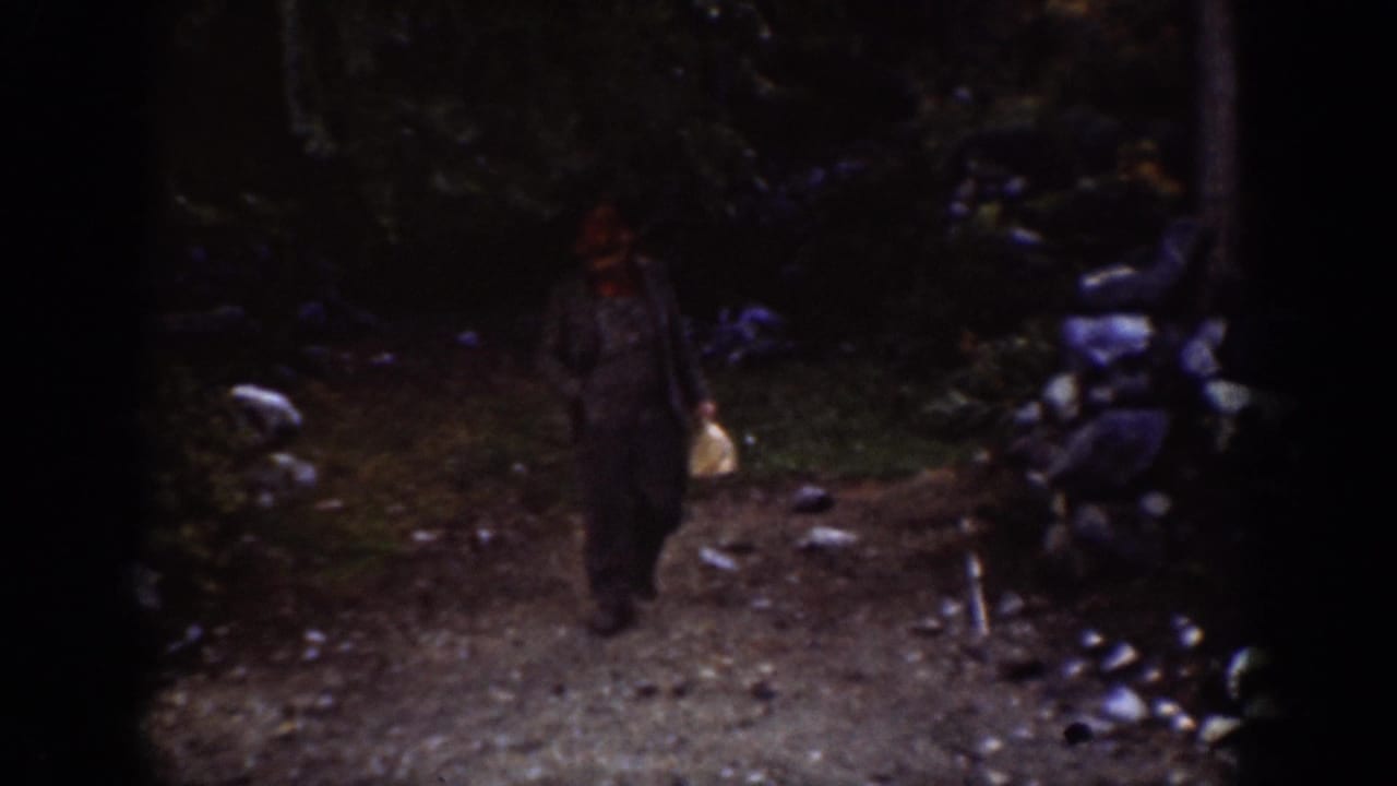 Single figure in a red hat walking toward camera on a gravel path with rocky terrain and trees.