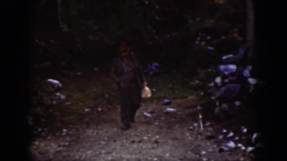 Adult male in red hat approaches along a rocky gravel path in a forested outdoor setting, consistent with 1930s Pacific Northwest travel footage.
