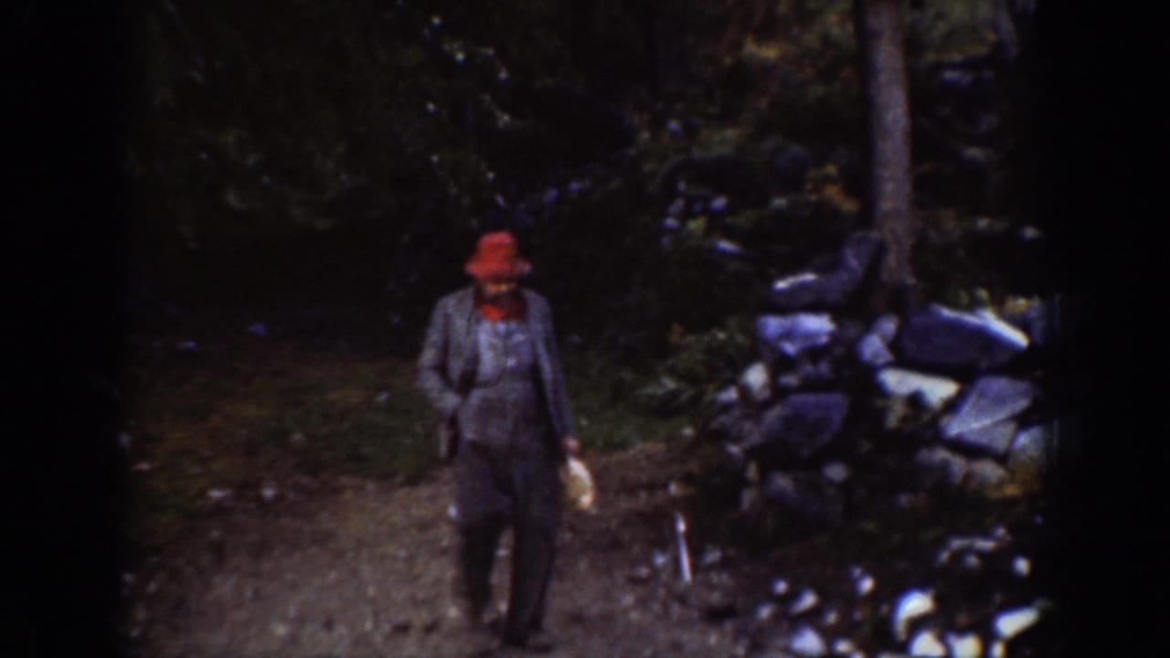 Adult male in red broad-brimmed hat and overalls walks a dirt path flanked by a dry-stone wall and dense conifers, circa 1939, Chelan, Washington.