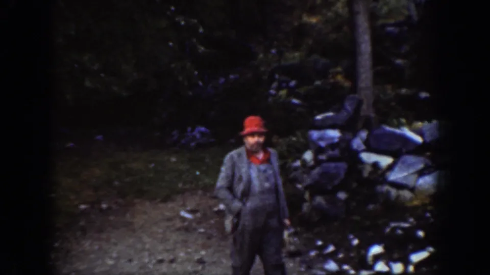 Adult male in red hat faces camera beside a dry-stone wall in a forested Pacific Northwest setting, circa 1939, Chelan, Washington.