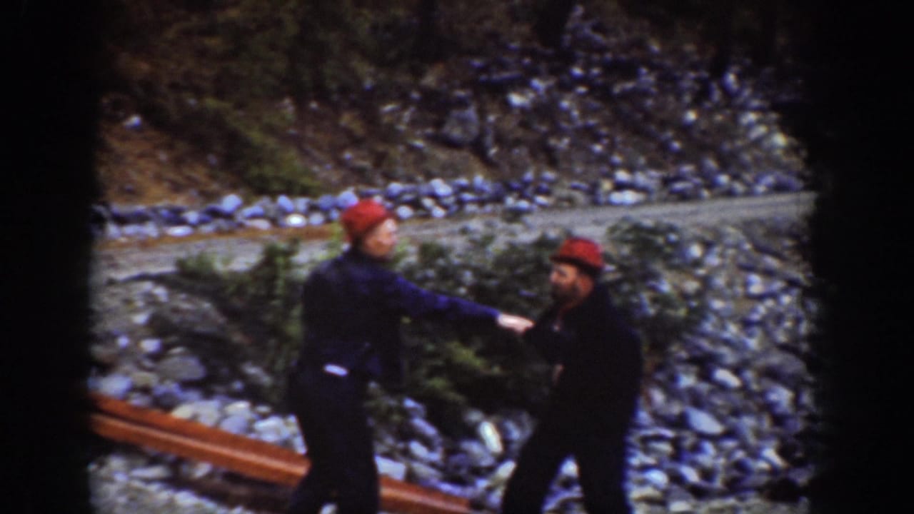 A man reaches or strikes toward another on a rocky Pacific Northwest lakeshore; both in red caps and dark coveralls, Chelan area, circa 1939.