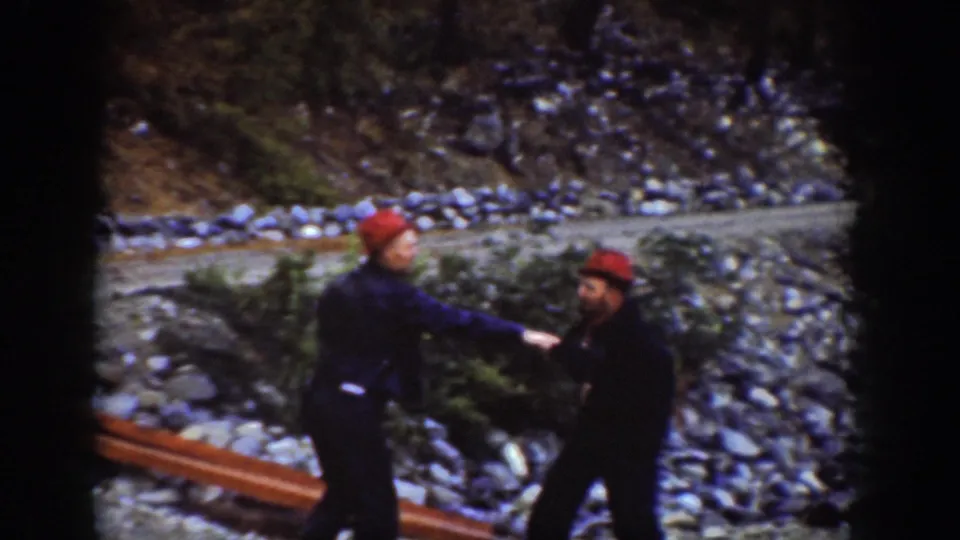 A man reaches or strikes toward another on a rocky Pacific Northwest lakeshore; both in red caps and dark coveralls, Chelan area, circa 1939.