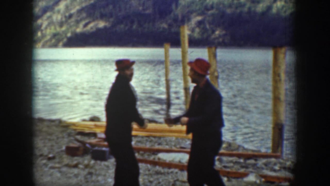 Two men in dark clothing and red caps face each other on a rocky lakeshore, with tall wooden posts and calm water behind them.