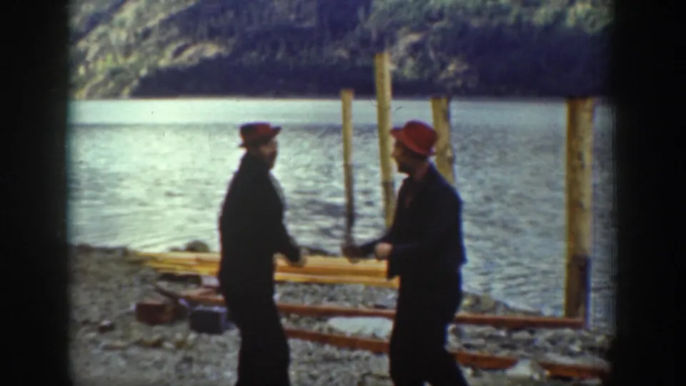 Two workers in red caps stand on the rocky shore of what appears to be Lake Chelan, Washington, circa 1939, with tall wooden pilings and forested mountains behind.