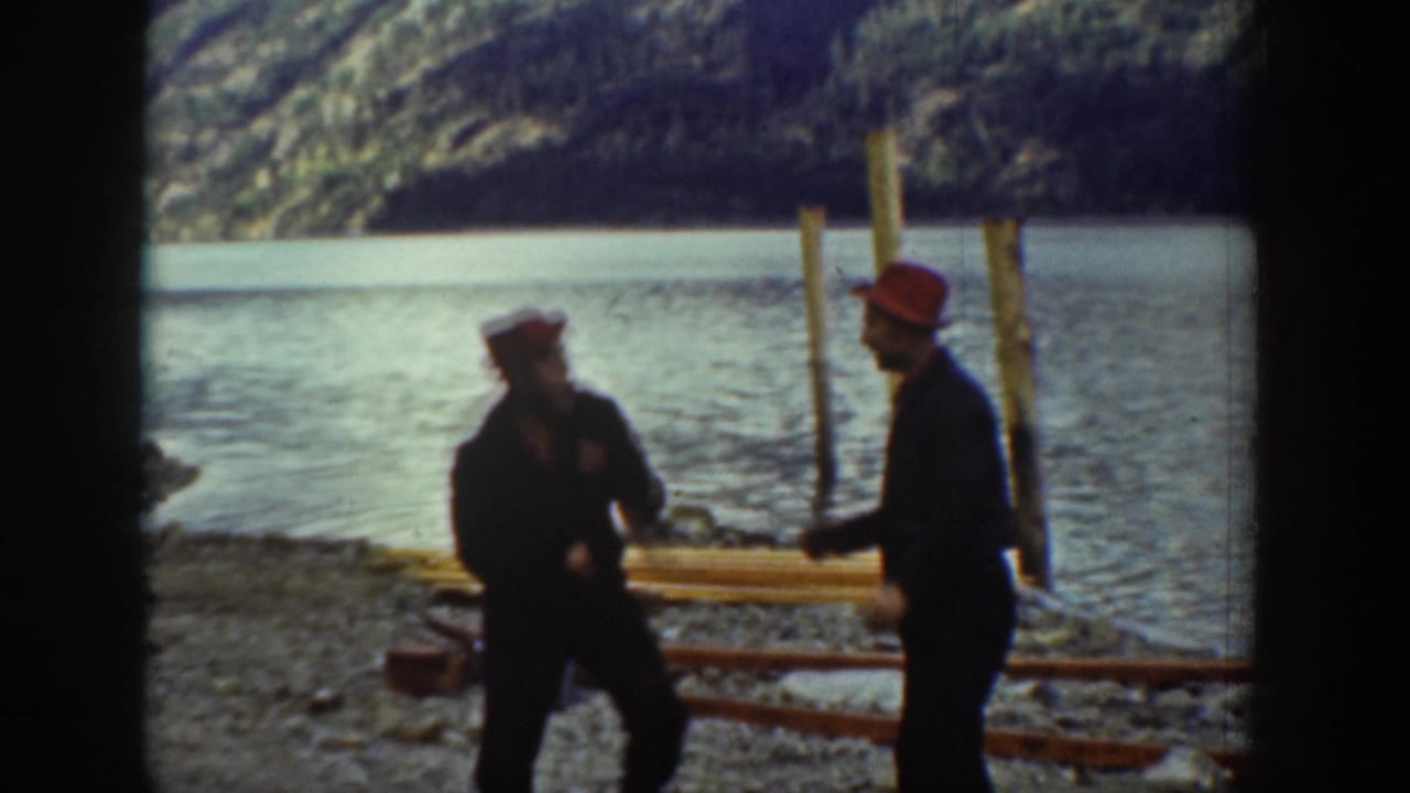 Two men in red work caps gesture toward each other on a rocky lakeshore in what may be Chelan, Washington, circa 1939, with wooden pilings and calm water in the background.