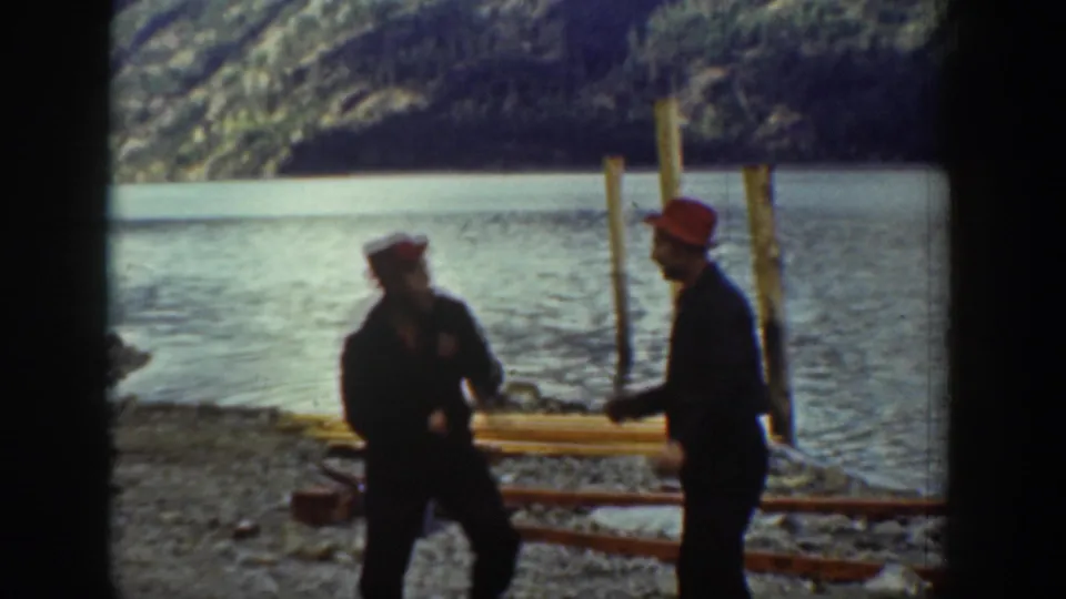 Two men in red work caps gesture toward each other on a rocky lakeshore in what may be Chelan, Washington, circa 1939, with wooden pilings and calm water in the background.