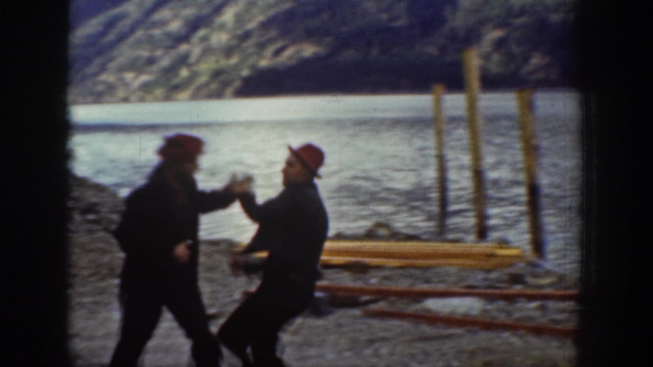 Two workers in red caps animate a discussion on a rocky Chelan-area lakeshore, circa 1939, with tall wooden pilings and a forested mountain slope in the background.