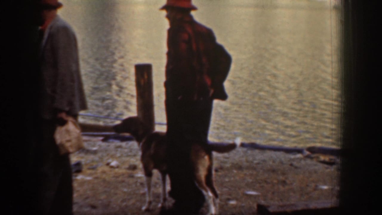 Two men in work jackets near a pier piling on a rocky shore, likely Lake Chelan, Washington, circa 1939; a dog stands in the foreground.
